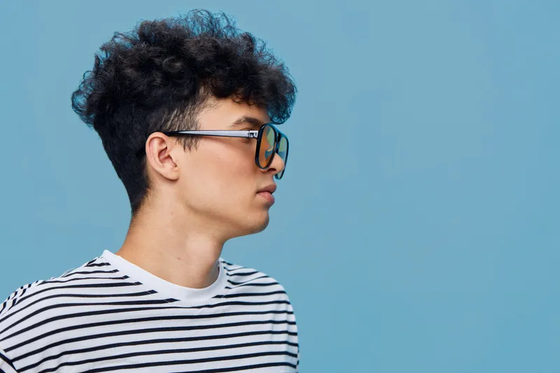 Young man with glasses wearing striped shirt looking sideways against blue background, showcasing casual style and modern fashion in a clean minimalist setting.