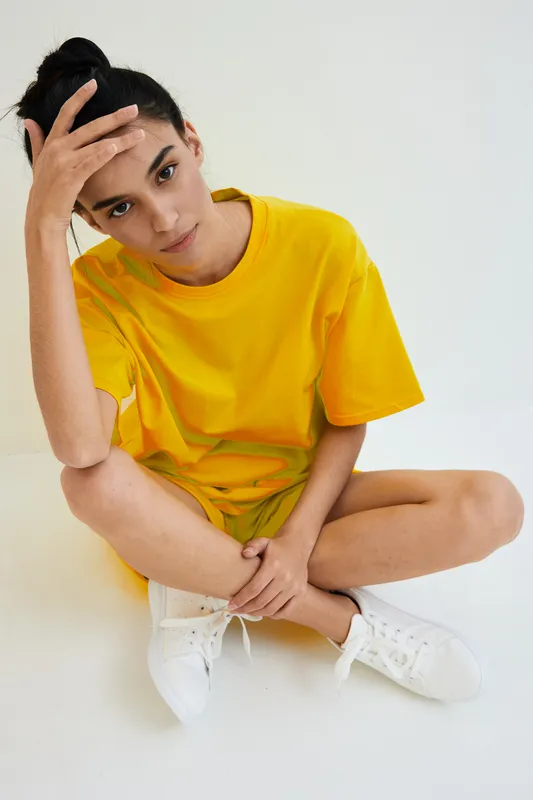 young woman in yellow t-shirt sitting cross-legged on white background, casual style, thoughtful expression, indoor minimalistic setting
