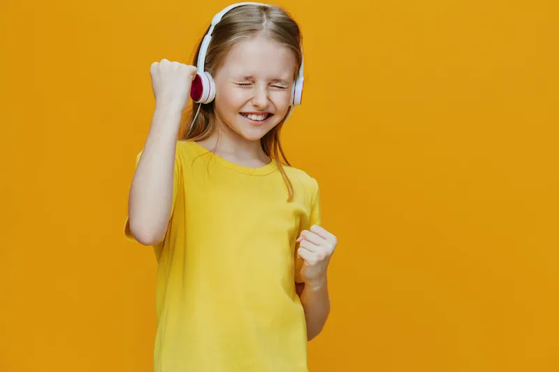 Happy girl in yellow shirt listening to music with headphones, celebrating joyfully against a vibrant orange background
