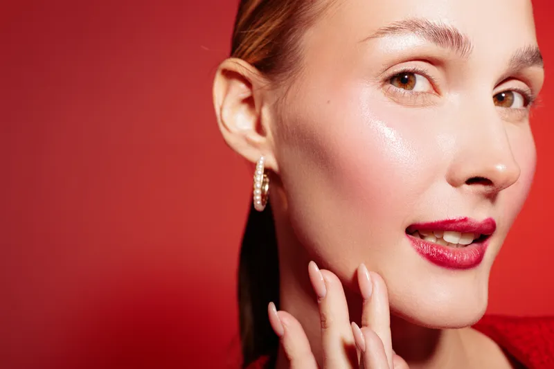 Close-up of a young woman with glowing skin and red lipstick touching her face. The red background and hoop earrings create a bold, stylish beauty portrait with confident expression.