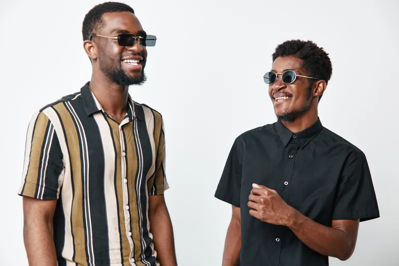 Two African American men with dark skin wearing sunglasses smiling and standing together in casual shirts against white background. Friendship and positive mood concept.