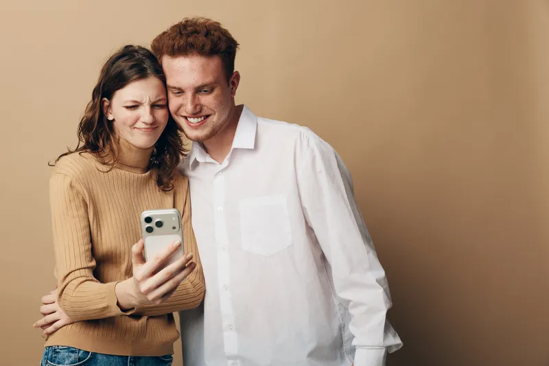 Young couple taking a selfie together with a smartphone, smiling and embracing each other against a plain beige background. Concept of love, friendship, and modern technology.