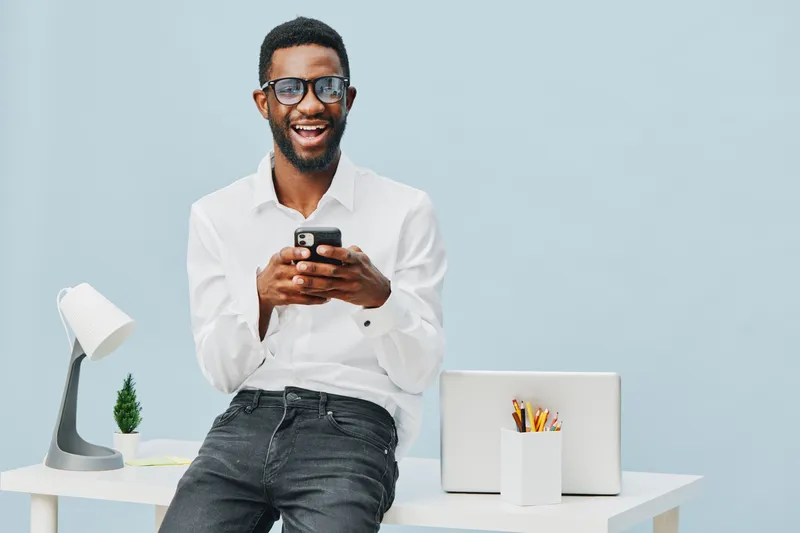 Smiling man using mobile phone while sitting at modern workspace with desk lamp, plants, and laptop, cheerful expression, casual style, indoor office environment, bright lighting, blue background, tec
