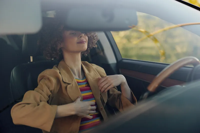 A stylish woman sits in a vintage car interior wearing a brown blazer over a striped top, adjusting her blazer with a calm confident expression in warm retro film tones.