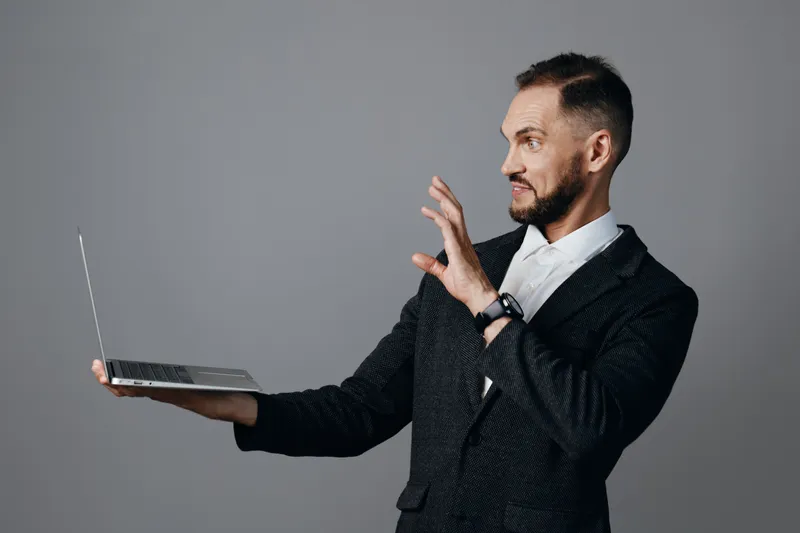 A professional businessman stands against a solid colored background, projecting confident emotion as he presents with a laptop held in one hand and gesturing.