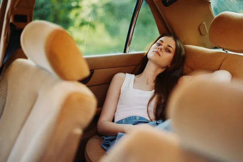 A young woman relaxing in a vintage car, dressed casually in a tank top and denim shorts, with a serene expression in a natural green setting.