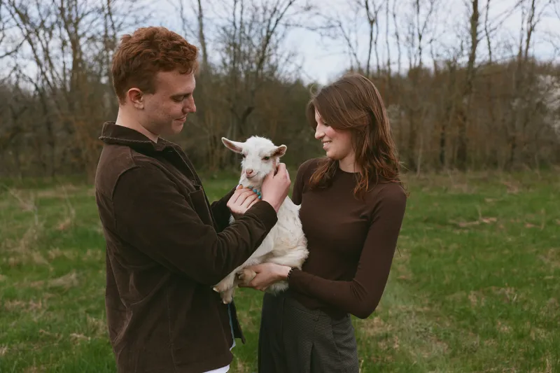 Cheerful couple outdoors, gentle interaction with a playful goat kid, warm smiles, soft breeze, natural colors, rustic field backdrop, casual jackets, candid moment of companionship