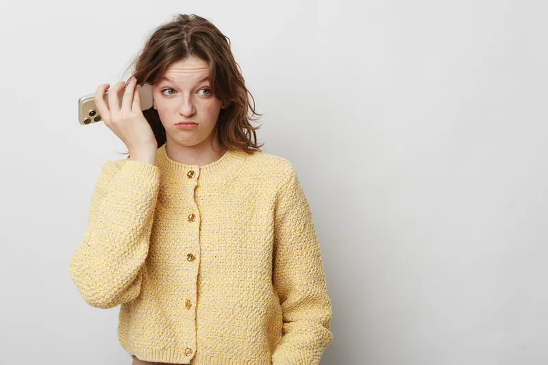 Young woman in yellow sweater holds smartphone to ear with confused expression, isolated on white background. Concept for misunderstanding, communication, and frustration in daily life.