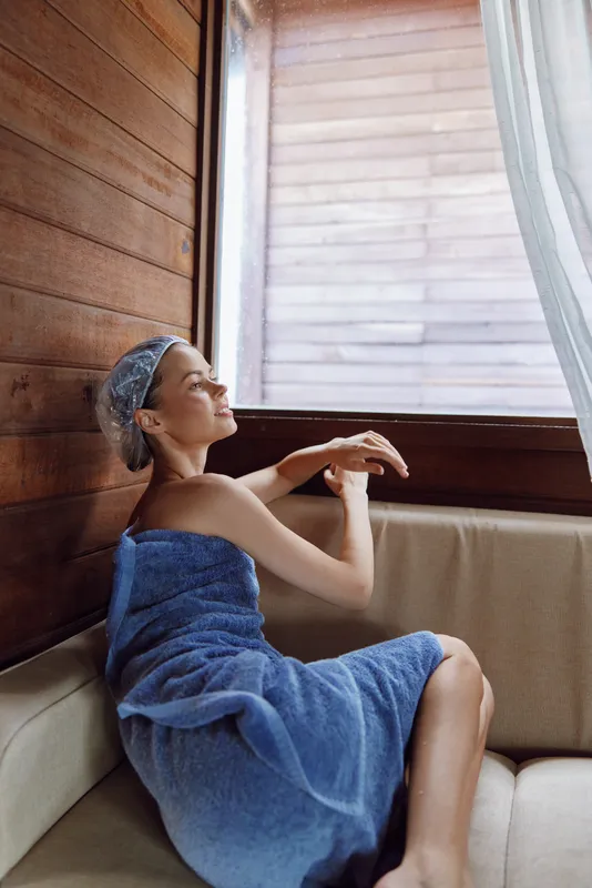 Relaxed woman wrapped in a blue towel and shower cap sitting on a beige sofa near a large window in a wooden interior, enjoying natural light and calm atmosphere.