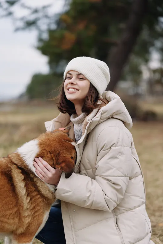 Woman in a warm beige coat and knit beanie hugs a fluffy dog outdoors, smiling softly in a chilly field with trees in the background.