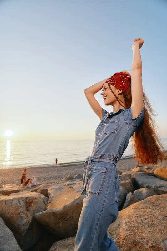 lifestyle woman in modern boho-western denim street style enjoys sunset on rocky beach with film color effect, carefree and relaxed mood, natural light