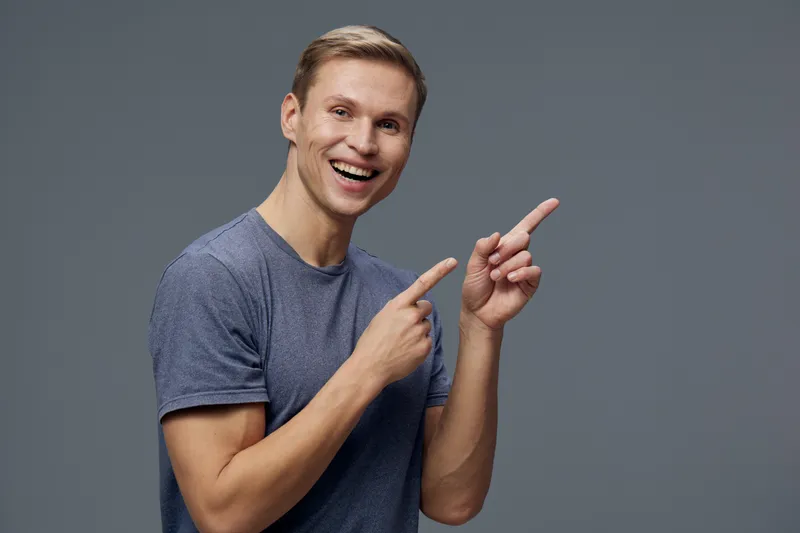 Smiling man with short blonde hair in casual blue t-shirt pointing to the side against plain gray background, expressing happiness and enthusiasm. Studio portrait. People lifestyle concept, positive e