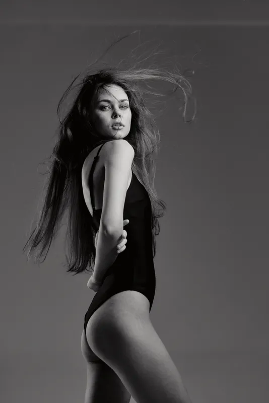 Black and white portrait of a young woman with long hair in a black bodysuit striking a confident pose, showcasing elegance and style.