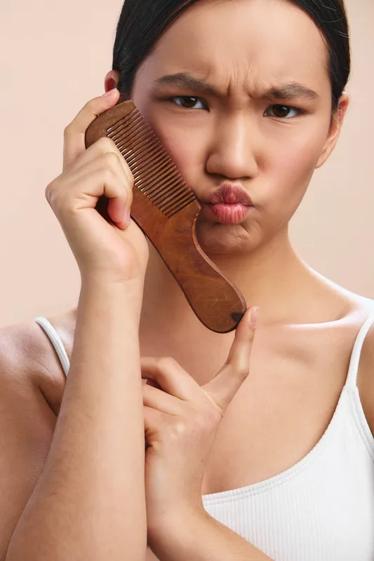 asian woman with a thoughtful pout as she holds a wide comb near her face, creating a candid grooming moment in a soft studio setting.