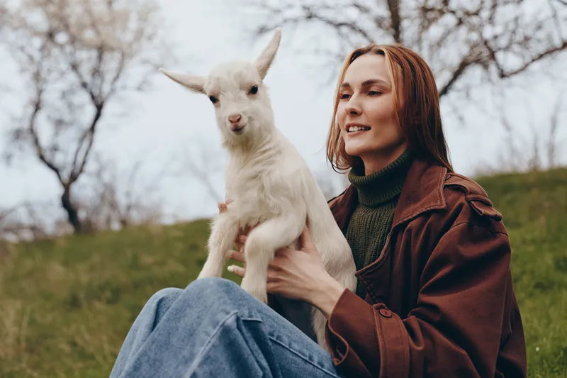 Goat, kid, woman, outdoors, lifestyle, nature, countryside, interaction, smiling, moment, woman sits on grass holding a young goat in a sunlit park, casual clothing, warm tones, relaxed vibe