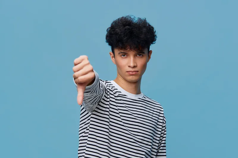 young man showing thumbs down gesture wearing striped shirt against blue background, expressing disapproval and dissatisfaction with serious facial expression.