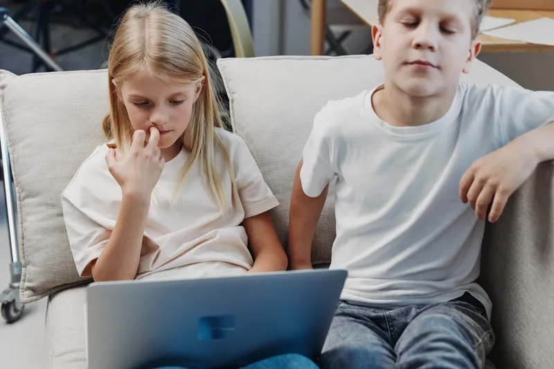 Two children sitting on a couch using a tablet, focused girl touching her nose and boy relaxing beside her. Concept of childhood, technology, leisure, and digital learning at home.
