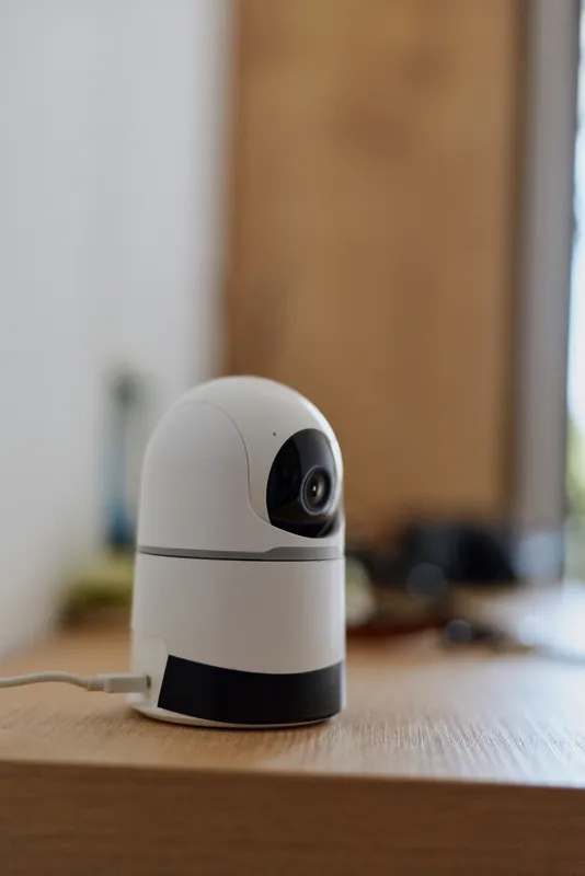 A small modern security camera sits on a wooden table in a bright home setting, suggesting monitoring and safety. Concept for home surveillance, privacy, and everyday tech use in daily life.