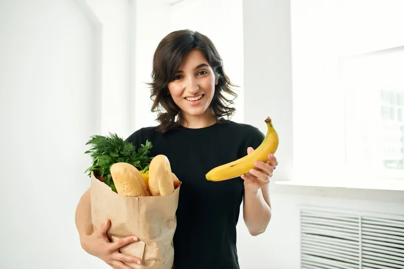 На фотографии запечатлена жизнерадостная женщина с пакетом продуктов, которая занимается домашними делами, связанными со здоровой пищей