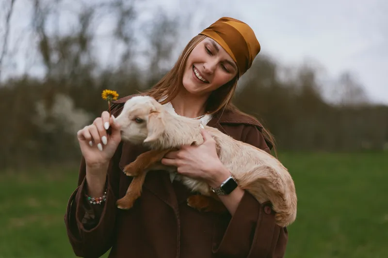 Young woman in brown coat and headscarf holds a baby goat gently while smiling outdoors in a green field during a calm spring day.