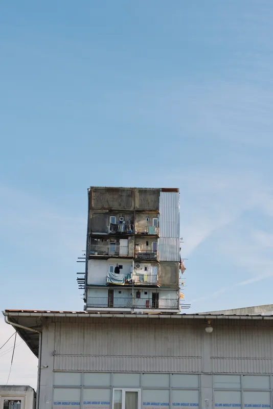 Tall stacked apartment tower atop a low industrial building under a clear blue sky, with small figures on balconies, urban housing structure and exposed living spaces.