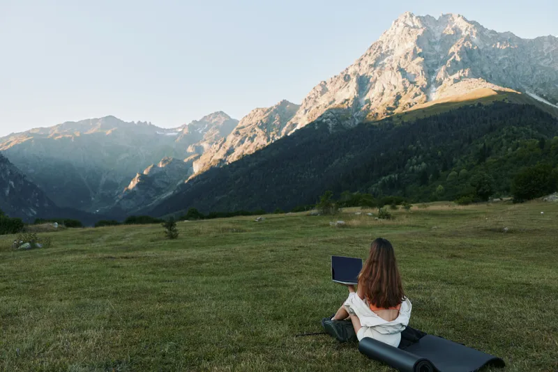 A lone person sits on a grassy meadow with a laptop, gazing toward a dramatic mountain range under a clear blue sky, enjoying peaceful remote work amid nature and fresh air.