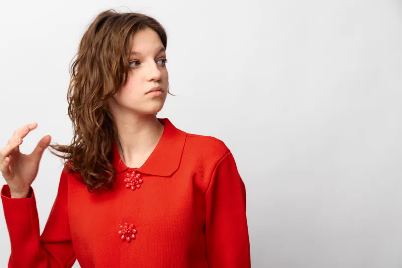Young woman in red sweater looks to the side with serious expression, touching her curly hair. Isolated on white background, concept of contemplation, fashion, and natural beauty.