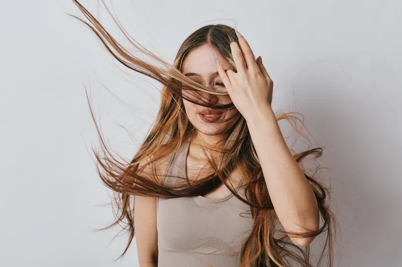 Young woman with long hair covering face using hand standing against plain background in casual tank top. Stylish female portrait with natural expression and dynamic hair movement indoors.