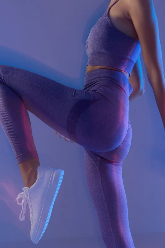 Woman sport fitness wearing purple activewear, stretching her leg against a blue glowing background, showcasing strength and flexibility in dynamic pose.