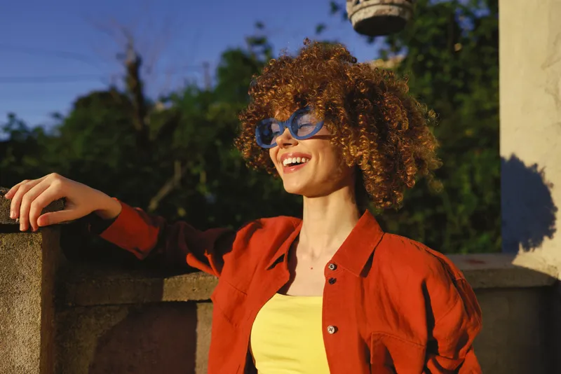 Young woman with curly hair smiles in bright sun, leaning on a stone railing in a warm outdoor setting, wearing a yellow top and orange shirt, radiating relaxed confidence