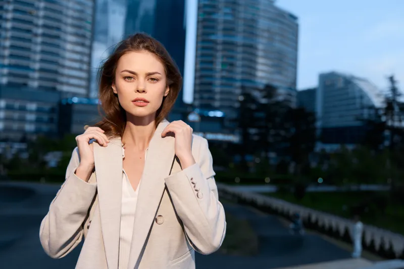 Young European woman in beige coat standing confidently in modern urban business district during daytime with office buildings background and clear sky