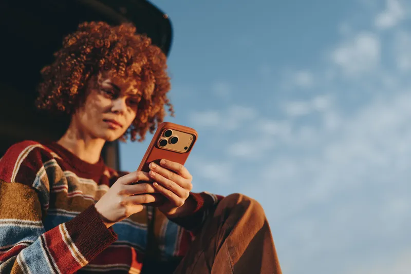 Woman with curly hair holding a smartphone outdoors, casual lifestyle portrait against blue sky, natural light, relaxed expression, warm plaid sweater, space for text