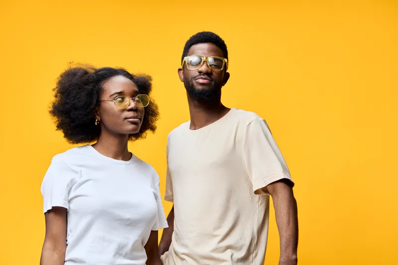 Two people with sunglasses posing in front of a vibrant yellow background, showcasing stylish summer wear and cheerful expressions for a contemporary lifestyle shoot