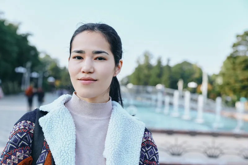 Woman outdoors in a park wearing a sweater and jacket, serene and casual, fashion lifestyle vibe with trees and water in the background, calm expression and relaxed street scene capturing a modern