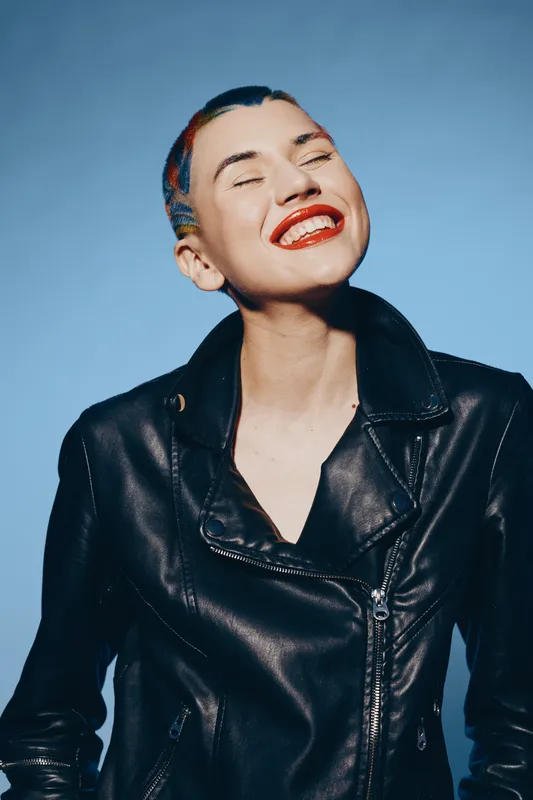 Young woman smiling brightly with colorful hair and red lipstick wearing a black leather jacket against a blue background, exuding bold and joyful energy.