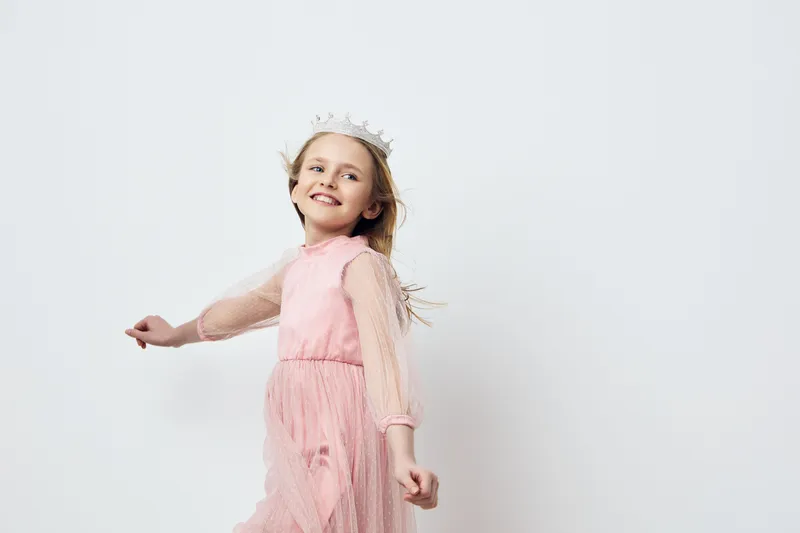 Cheerful girl in a pink dress with a crown, joyfully twirling against a white background, embodying childhood happiness and innocence