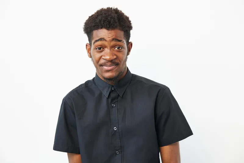 A smiling black man wearing a casual black shirt, exuding charm and warmth while looking directly at the camera. Emotional connection in a contemporary setting.