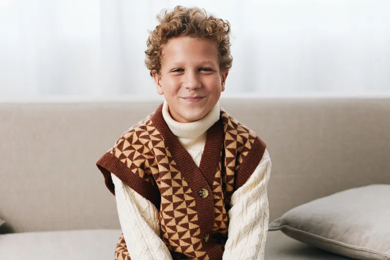 A smiling young boy sits on a sofa wearing a patterned knit vest over a cream turtleneck, creating a cozy, vintage inspired portrait with warm natural light.