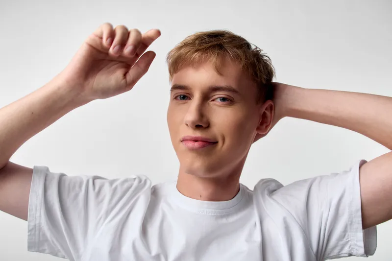 Casual young man in a white t shirt, arms raised behind his head, relaxed smile, studio lighting, clean background, everyday lifestyle mood and confident posture.