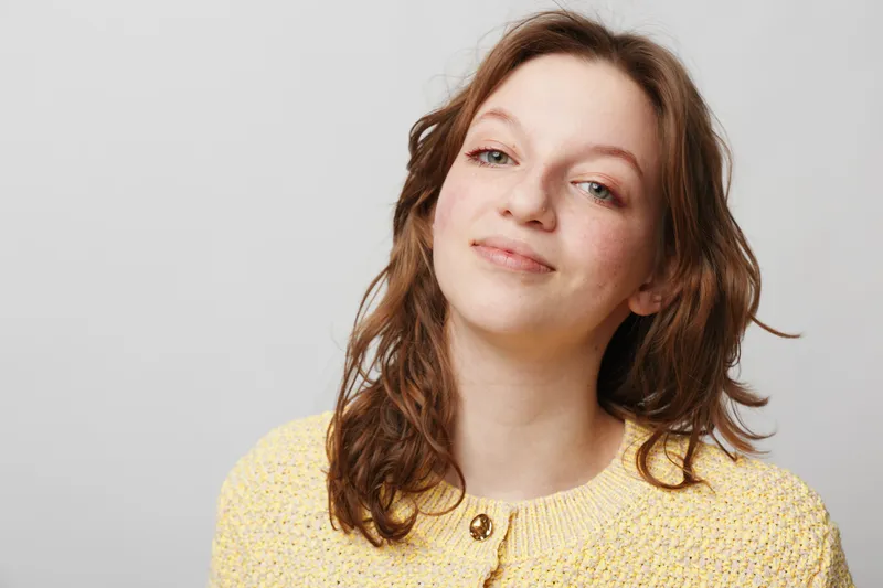 Young woman with wavy brown hair wearing yellow sweater smiles gently against light background. Portrait of confident female with natural makeup and relaxed expression in studio setting.