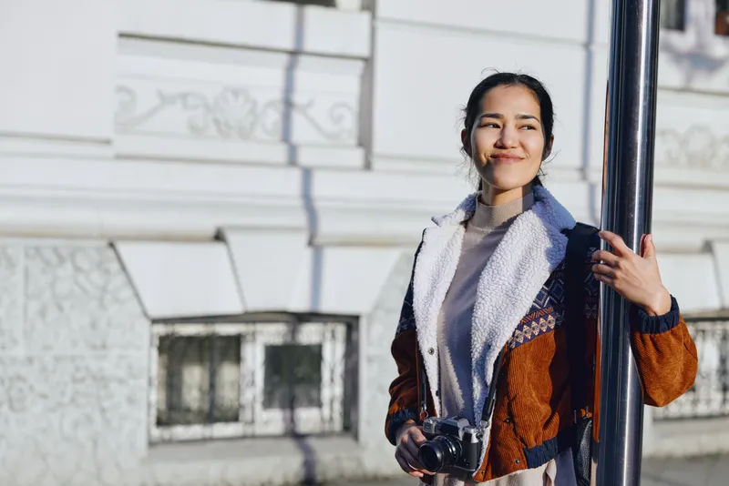 Woman with camera and warm jacket stands beside a pole in an urban facade, enjoying outdoor photography and a candid sunny moment in a city street scene, natural light