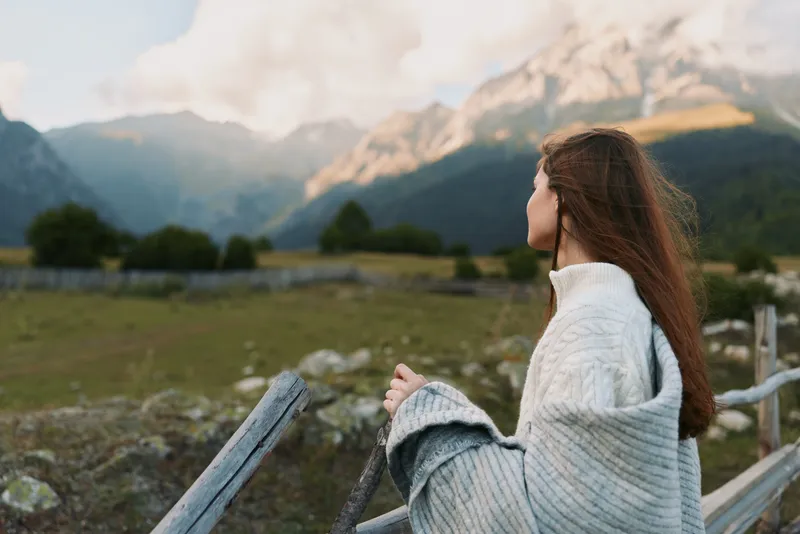 A young woman wearing a grey cardigan stands by a wooden railing, gazing toward distant mountains with a calm, contemplative mood under soft natural light, creating a peaceful, scenic moment.