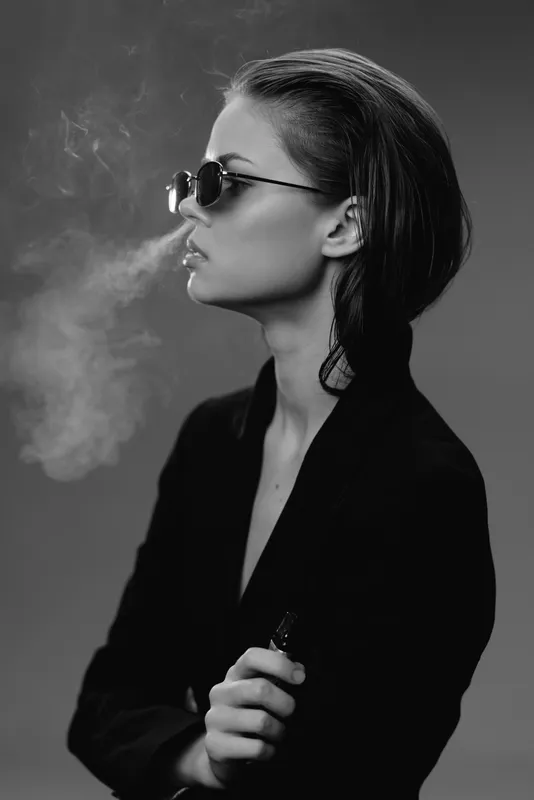 Black and white portrait of a stylish woman wearing sunglasses and smoking with smoke around her face.