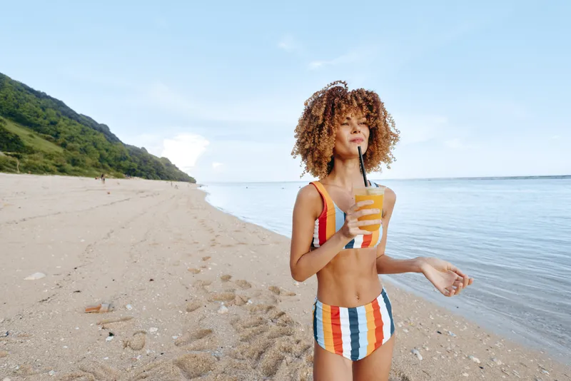 Woman on beach by seaside ocean in summer, holding snack package with curly hair, wearing swimsuit, sunny outdoor portrait, relaxed vacation lifestyle, fun eating concept.