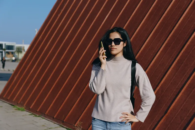 Sunglasses, woman on phone outdoors beside a diagonal rust wall, casual fashion pose, stylish setting, sunny day, street style influence, contemporary accessory, confident pose in an urban landscape