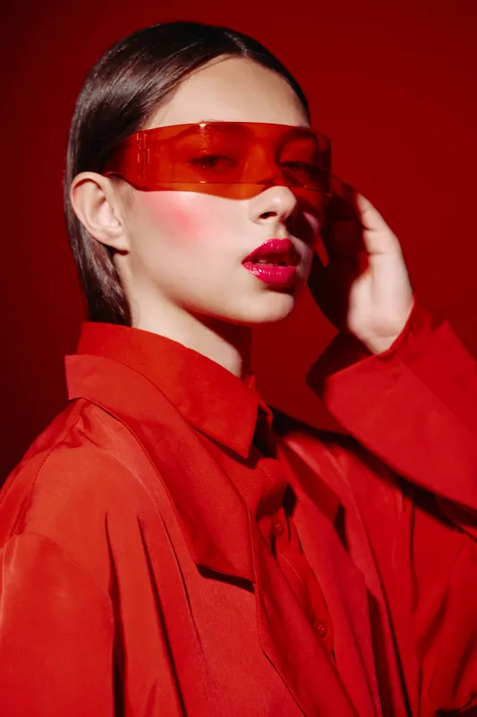 Fashion model in oversized red suit and translucent red visor poses against red background with bright red lips and graphic orange eyeshadow. Editorial high fashion concept.