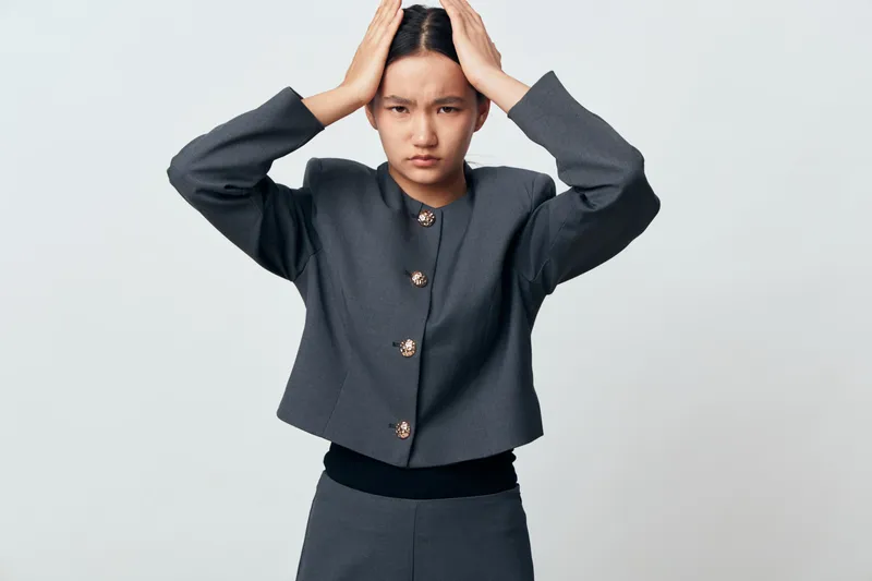 Woman standing with hands on head looking stressed and worried in gray jacket and pants. Young female with serious expression on light background in studio for mental health and pressure concept.
