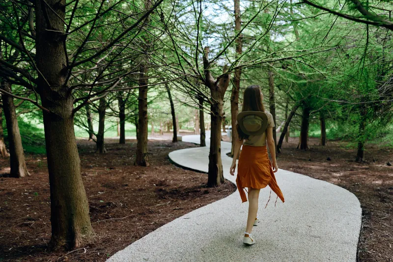 Woman walking on winding path through forest with backpack and hat, trees overhead and sunlight filtering, nature trail scene for travel, solitude and summer outdoor exploration.