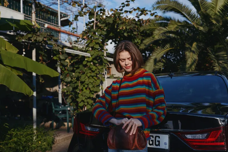 Woman leaning on car trunk wearing a colorful striped sweater and holding a brown leather bag, outdoor portrait in sunlight by palm trees and urban garden, casual fashion moment and relaxed pose