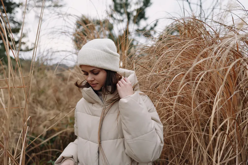 Winter woman outdoors wearing a beige coat and knit hat stands among tall grasses, creating a calm field scene. A cozy lifestyle portrait that highlights nature, warmth, and seasonal fashion in muted